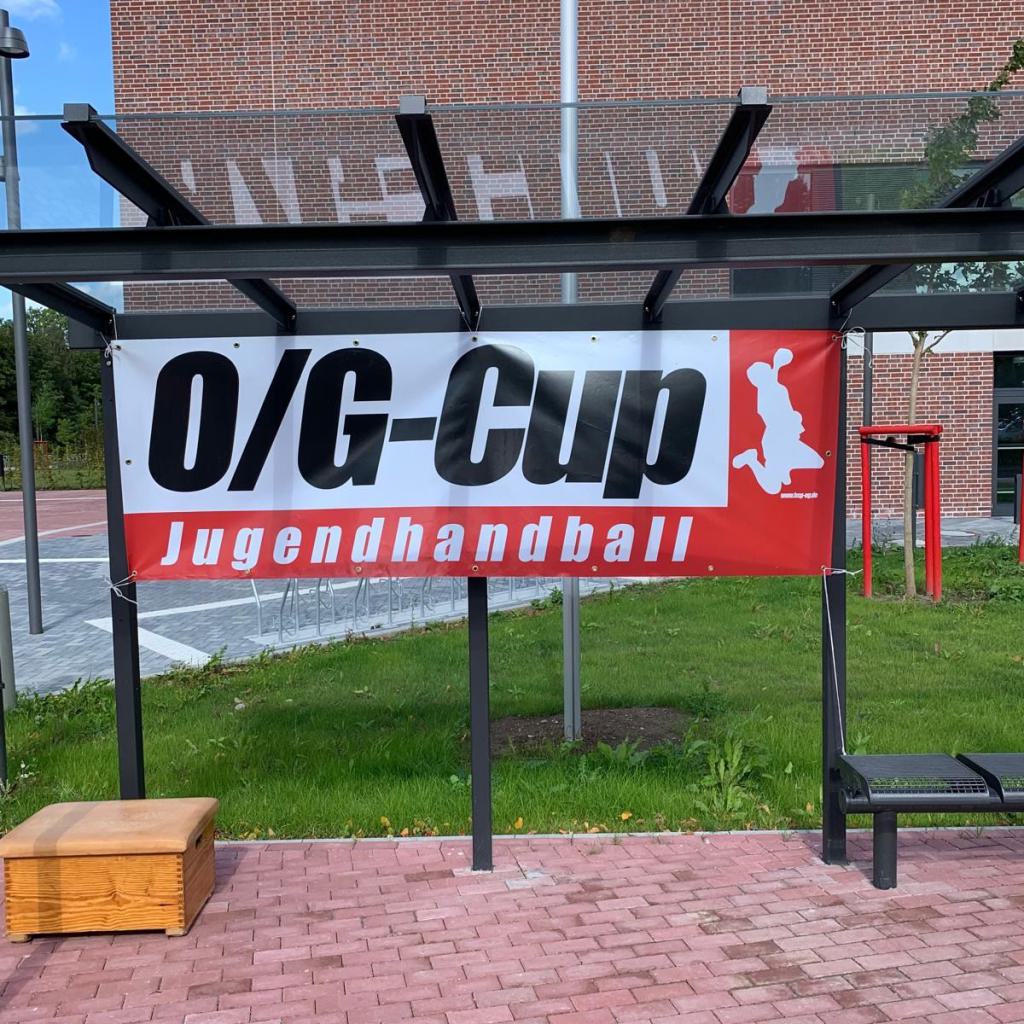 Banner mit der Aufschrift O/G-Cup Jugendhandball an einem überdachten Sitzbereich im Freien