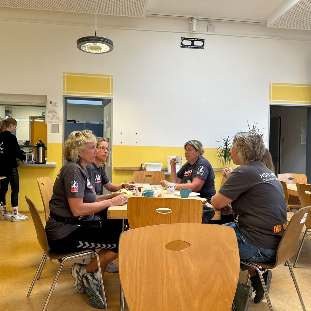 Mehrere Frauen sitzen an einem Tisch in einem Pausenraum und essen gemeinsam, im Hintergrund steht eine weitere Frau am Tresen.