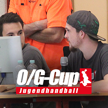 Vier junge Männer sitzen an einem Tisch mit Computern und Mikrofonen, darunter ein Mann in orangem Shirt.