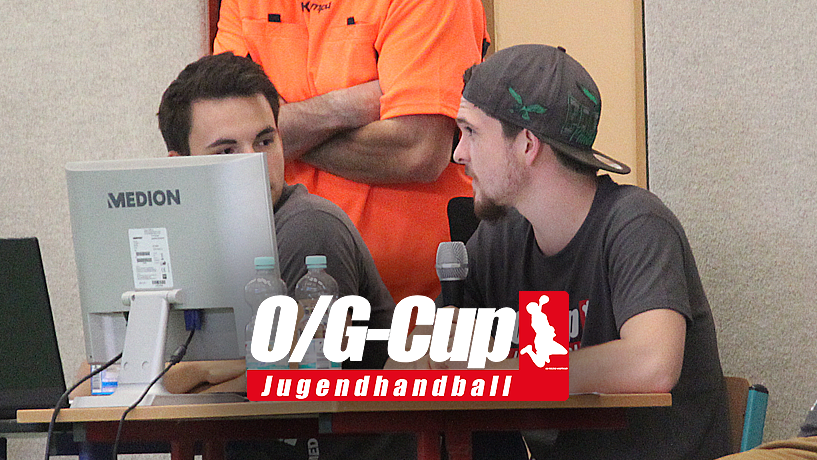 Vier junge Männer sitzen an einem Tisch mit Computern und Mikrofonen, darunter ein Mann in orangem Shirt.