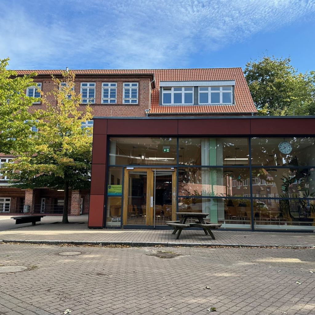 Schulgebäude mit Bänken und Bäumen vor Eingangsbereich mit großen Fenstern